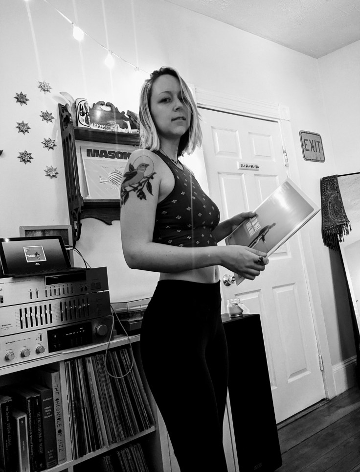 A black-and-white image of a female holding a vinyl record and standing in front of a stereo system